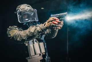 A person in tactical gear wearing a clear face shield and aiming a handgun, with smoke and a bright muzzle flash against a dark background.